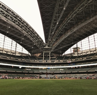 An expansive sports stadium with a large, open field of lush green grass and a partially retractable roof, showcasing a vast seating area surrounding the field. A group of people is visible along the boundary, seemingly engaged in an event or tour.