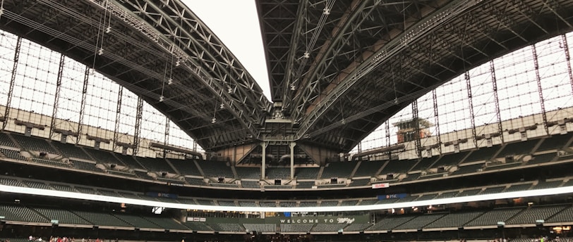 An expansive sports stadium with a large, open field of lush green grass and a partially retractable roof, showcasing a vast seating area surrounding the field. A group of people is visible along the boundary, seemingly engaged in an event or tour.