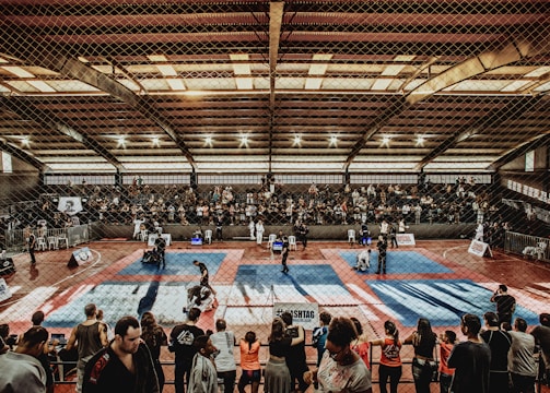 A large indoor arena with a high ceiling, filled with an audience seated on bleachers. In the center, two fighters are engaged in a martial arts competition on blue and red mats. The scene is captured through a mesh fence, adding a textured foreground.