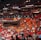 An indoor sports arena filled with a large crowd of spectators. People are sitting in tiered seating areas with red and orange tones dominating the seats. A person in the foreground is waving a large white flag with 'MIAMI' written on it. Numerous attendees wear basketball jerseys, and there is visible excitement and energy in the atmosphere. Advertisements for Uber are displayed prominently on digital boards near the ceiling.