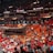 An indoor sports arena filled with a large crowd of spectators. People are sitting in tiered seating areas with red and orange tones dominating the seats. A person in the foreground is waving a large white flag with 'MIAMI' written on it. Numerous attendees wear basketball jerseys, and there is visible excitement and energy in the atmosphere. Advertisements for Uber are displayed prominently on digital boards near the ceiling.