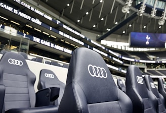 A row of black leather stadium seats with the Audi logo prominently displayed in an upscale sports venue. In the background, there are large screens and bright signage displaying the words 'The game is about glory,' suggesting a high-profile sporting event location.