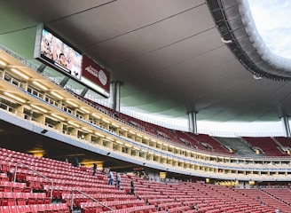 A large, modern stadium with red seats and a sleek, expansive roof structure. A few people are scattered among the seats, and there are large digital screens displaying advertisements. The setting is well-lit with multiple levels visible, suggesting a multi-tiered structure.