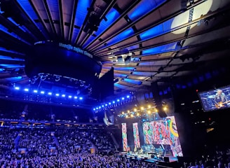 A large indoor arena filled with a crowd attending a live concert. Bright stage lights illuminate the scene, with colorful vertical screens displaying vibrant patterns. The ceiling showcases architectural beams, most prominently lit in blue hues.