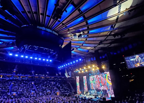 A large indoor arena filled with a crowd attending a live concert. Bright stage lights illuminate the scene, with colorful vertical screens displaying vibrant patterns. The ceiling showcases architectural beams, most prominently lit in blue hues.