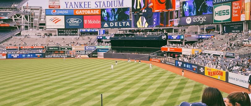 A baseball stadium filled with spectators watching a game on a neatly maintained grass field. The stadium features large advertising screens displaying various logos and brands. Fans sitting in the foreground are dressed casually, enjoying the sporting event.