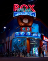 A vibrant, neon-lit facade of a gaming and sports establishment at night, featuring a large glove holding a baseball above the entrance. The building is adorned with colorful signs in both English and Japanese, with various advertisements and an illuminated sign that says 'ROX DOME'. Several people are walking towards the entrance, and bicycles are parked along the side.