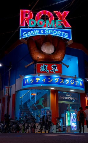 A vibrant, neon-lit facade of a gaming and sports establishment at night, featuring a large glove holding a baseball above the entrance. The building is adorned with colorful signs in both English and Japanese, with various advertisements and an illuminated sign that says 'ROX DOME'. Several people are walking towards the entrance, and bicycles are parked along the side.