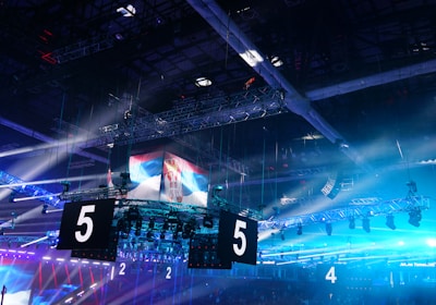 A large indoor arena is equipped with advanced lighting and audio systems. Suspended above the space are numerous digital displays showcasing numbers. The environment is filled with blue illumination and spotlights creating a vibrant atmosphere.