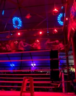 A brightly lit boxing or wrestling ring with vivid red and blue lights. Banners with images of fighters hang from the ceiling. The ropes of the ring are prominently visible, and a stool is placed inside the ring. The arena is filled with an audience, silhouetted against the colorful lighting.