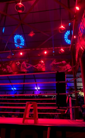 A brightly lit boxing or wrestling ring with vivid red and blue lights. Banners with images of fighters hang from the ceiling. The ropes of the ring are prominently visible, and a stool is placed inside the ring. The arena is filled with an audience, silhouetted against the colorful lighting.