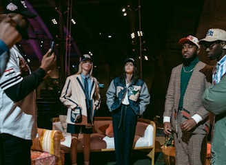 A group of people are gathered indoors, wearing stylish casual and sports-themed outfits, including jackets and caps. Some are standing while one appears to be taking a photo. The environment has a warm, wooden interior with visible beams and spotlights, and a cozy arrangement of furniture including chairs and a sofa.