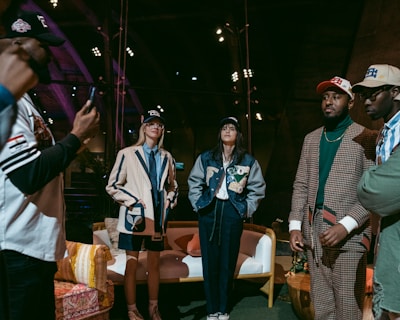 A group of people are gathered indoors, wearing stylish casual and sports-themed outfits, including jackets and caps. Some are standing while one appears to be taking a photo. The environment has a warm, wooden interior with visible beams and spotlights, and a cozy arrangement of furniture including chairs and a sofa.