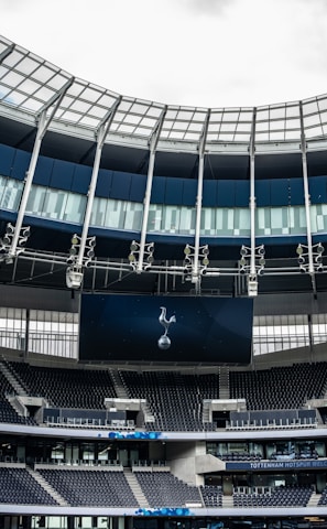 A modern stadium interior featuring a large digital screen with a logo prominently displayed. The seating areas are empty, with dark-colored seats and some structural elements like beams and glass walls visible. A combination of dark blue and neutral tones are prominent.