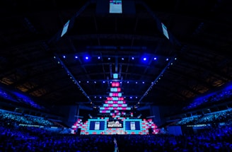 An expansive venue filled with a large audience, featuring a grand stage lit with vibrant blue and pink lights. The ceiling is decorated with a large display, and numerous screens showcase the event's branding. The atmosphere is dynamic and futuristic, reflecting a technologically advanced conference.