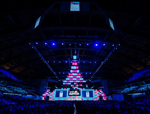 An expansive venue filled with a large audience, featuring a grand stage lit with vibrant blue and pink lights. The ceiling is decorated with a large display, and numerous screens showcase the event's branding. The atmosphere is dynamic and futuristic, reflecting a technologically advanced conference.