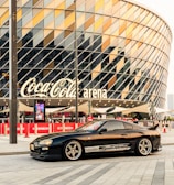 A sleek black sports car is parked in front of a modern arena with a distinctive geometric glass facade. The arena features a large sign with bold lettering. Paved area surrounds the car, creating an urban, contemporary setting.