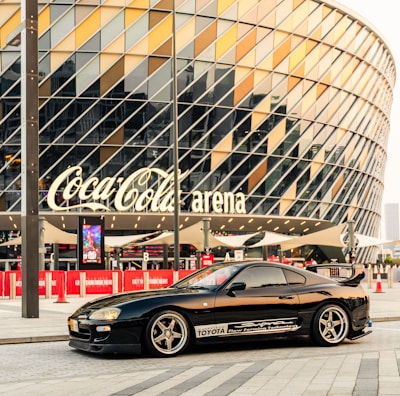 A sleek black sports car is parked in front of a modern arena with a distinctive geometric glass facade. The arena features a large sign with bold lettering. Paved area surrounds the car, creating an urban, contemporary setting.