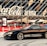 A sleek black sports car is parked in front of a modern arena with large glass panels and a prominent Coca-Cola sign. The car has customized decals and alloy wheels, and the scene is set in an urban environment with red banners and pavement.