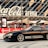 A sleek black sports car is parked in front of a modern arena with large glass panels and a prominent Coca-Cola sign. The car has customized decals and alloy wheels, and the scene is set in an urban environment with red banners and pavement.