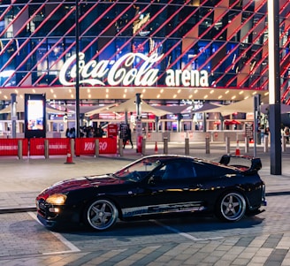 A sleek black sports car is parked in front of an illuminated arena entrance, with a large sign bearing the name of a famous beverage brand. The building features a modern, geometric facade with bright red and blue lights, creating a vibrant and lively atmosphere.